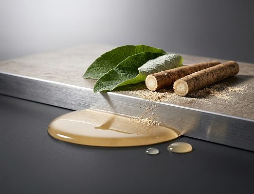 Raw jute twigs and green leaves with a pool of golden fiber extract on a dark surface, set against a brushed metal block.
