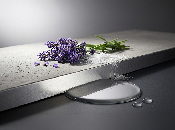 A close-up studio shot of a metallic silver surface with a clear liquid puddle, white fibers, and a small bunch of purple lavender on top, set against a dark gray background.