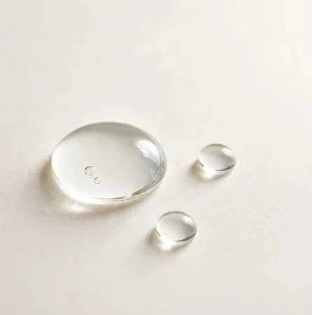 Three clear water droplets of varying sizes resting on a smooth, off-white surface, with the largest droplet showing subtle internal reflections.