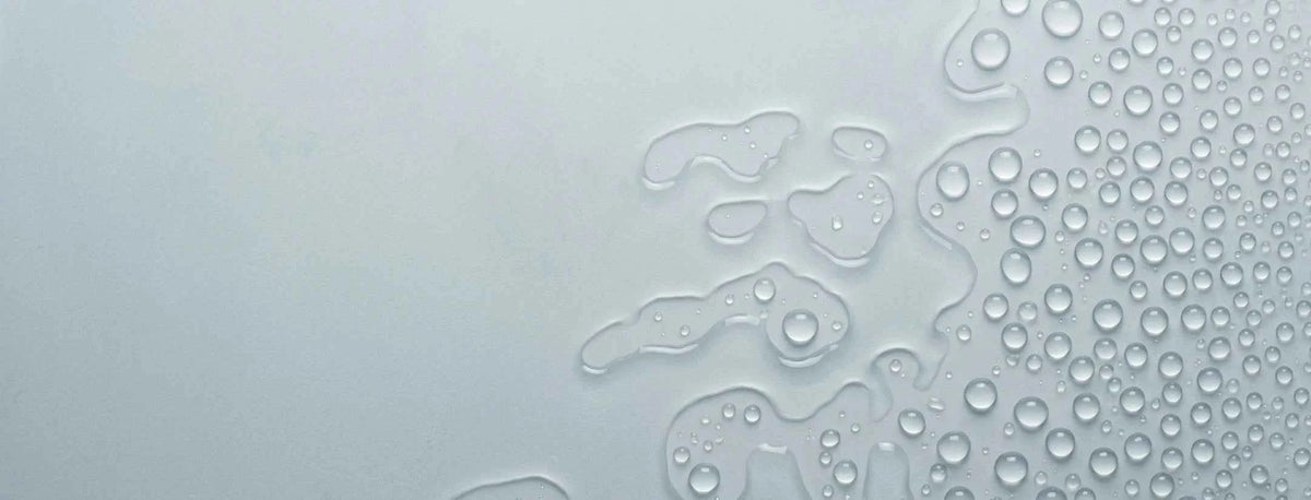 A close-up of a translucent, frosted surface with water droplets forming organic shapes across the right side against a pale gray background.