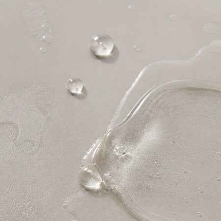 A close-up of a clear, glossy liquid or gel swatch on a light beige textured surface, with three distinct water droplets above it.