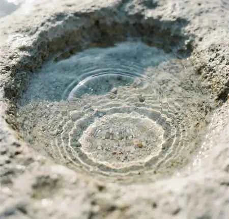 A close-up view of clear water pooling in a rough, sunlit stone or concrete basin, with concentric ripples spreading from the center.