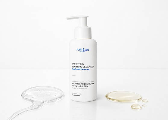 A white pump bottle of Ariège Purifying Foaming Cleanser stands on a white surface with a clear gel swab on the left and two gold-toned liquid droplets on the right, against a clean white background.