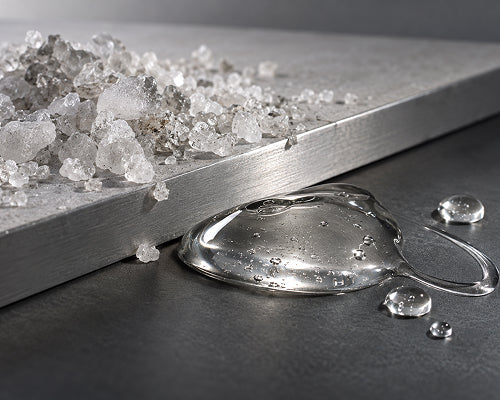 A close-up studio shot of a transparent gel or adhesive droplet on a dark surface, with a brushed metal board and scattered translucent crystal-like pieces behind it.