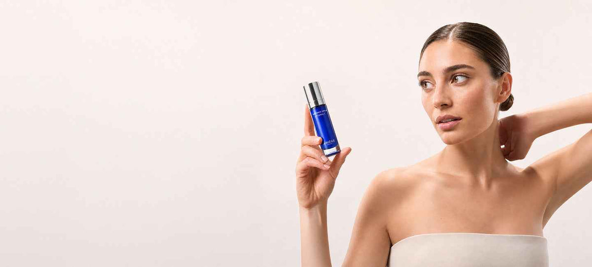 A woman with her hair pulled back wears a beige strapless top and holds up a blue cosmetic bottle with a silver cap against a plain light gray background.