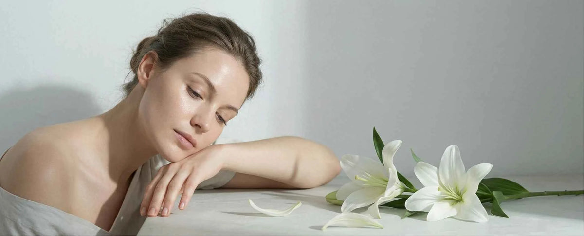 A woman rests her head on her folded arms on a white table, with white lilies and a few petals beside her in a softly lit, minimalist setting.