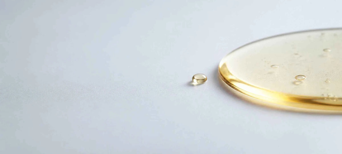 A close-up of a translucent golden liquid, likely an oil or serum, with a small droplet beside a shallow pool on a light gray surface.