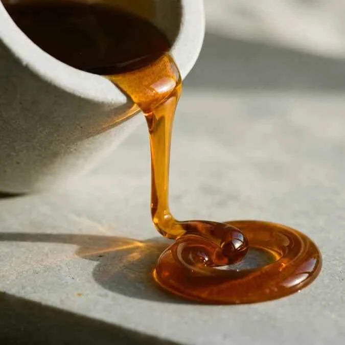 Thick, glossy amber honey or syrup being poured from a white ceramic pitcher onto a light stone surface, forming a curled pool of translucent golden-brown liquid.